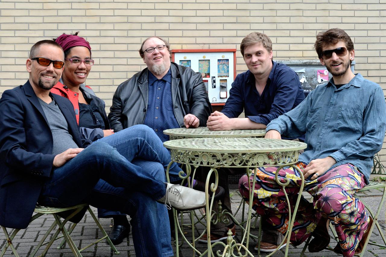 Die fünf Mitglieder der Musikergruppe sitzen um historisierende, grüne Metallgartentische und lachen in die Kamera.