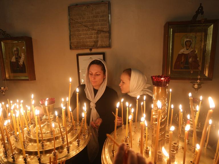 Zwei Frauen mit je einem Tuch über dem Kopf zünden während eines orthodoxen Gottesdienstes in der Kazaner Kathedrale in St. Petersburg Kerzen an, hinter ihnen sind Ikonen zu sehen.