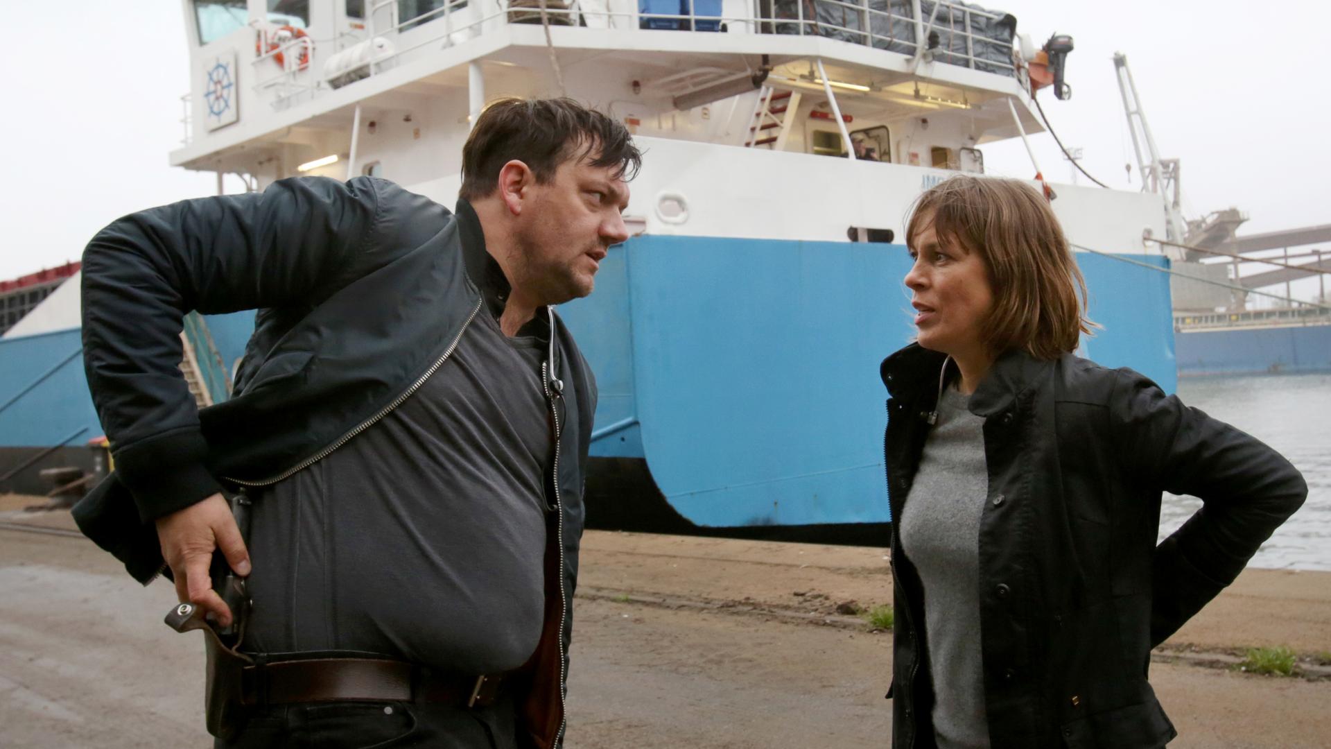 Ein Mann und eine Frau stehen vor einem Schiff im Hafen. Der Mann steckt sich eine Pistole in den Gürtel.
