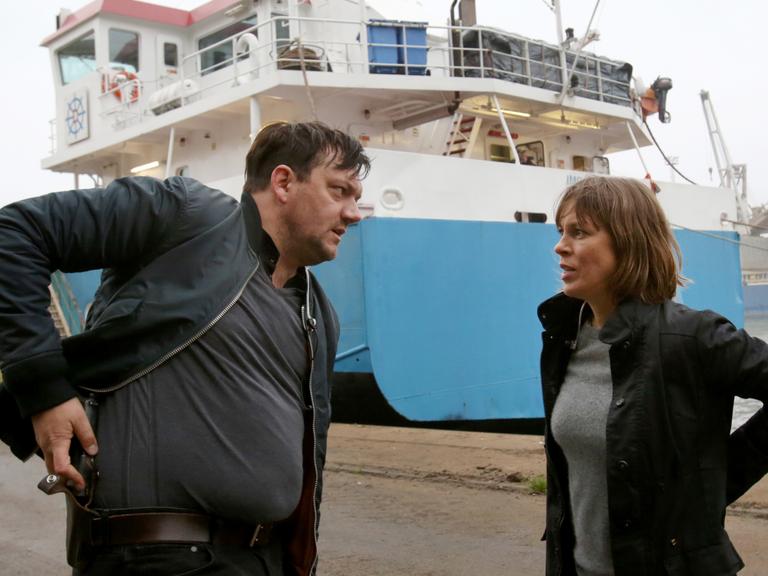 Ein Mann und eine Frau stehen vor einem Schiff im Hafen. Der Mann steckt sich eine Pistole in den Gürtel.