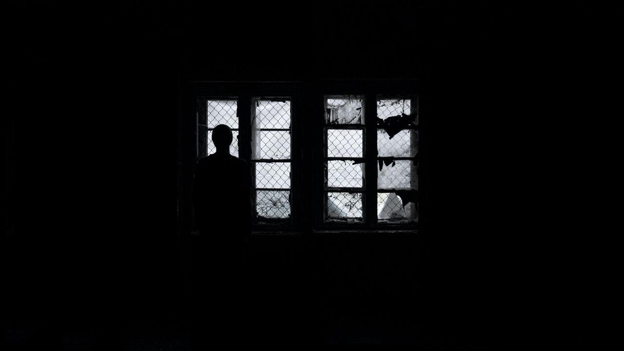Eine Frau steht als Silhouette im Dunkeln vor vergitterten Fenstern und schaut hinaus Eine Frau steht als Silhouette im Dunkeln vor vergitterten Fenstern und schaut hinaus