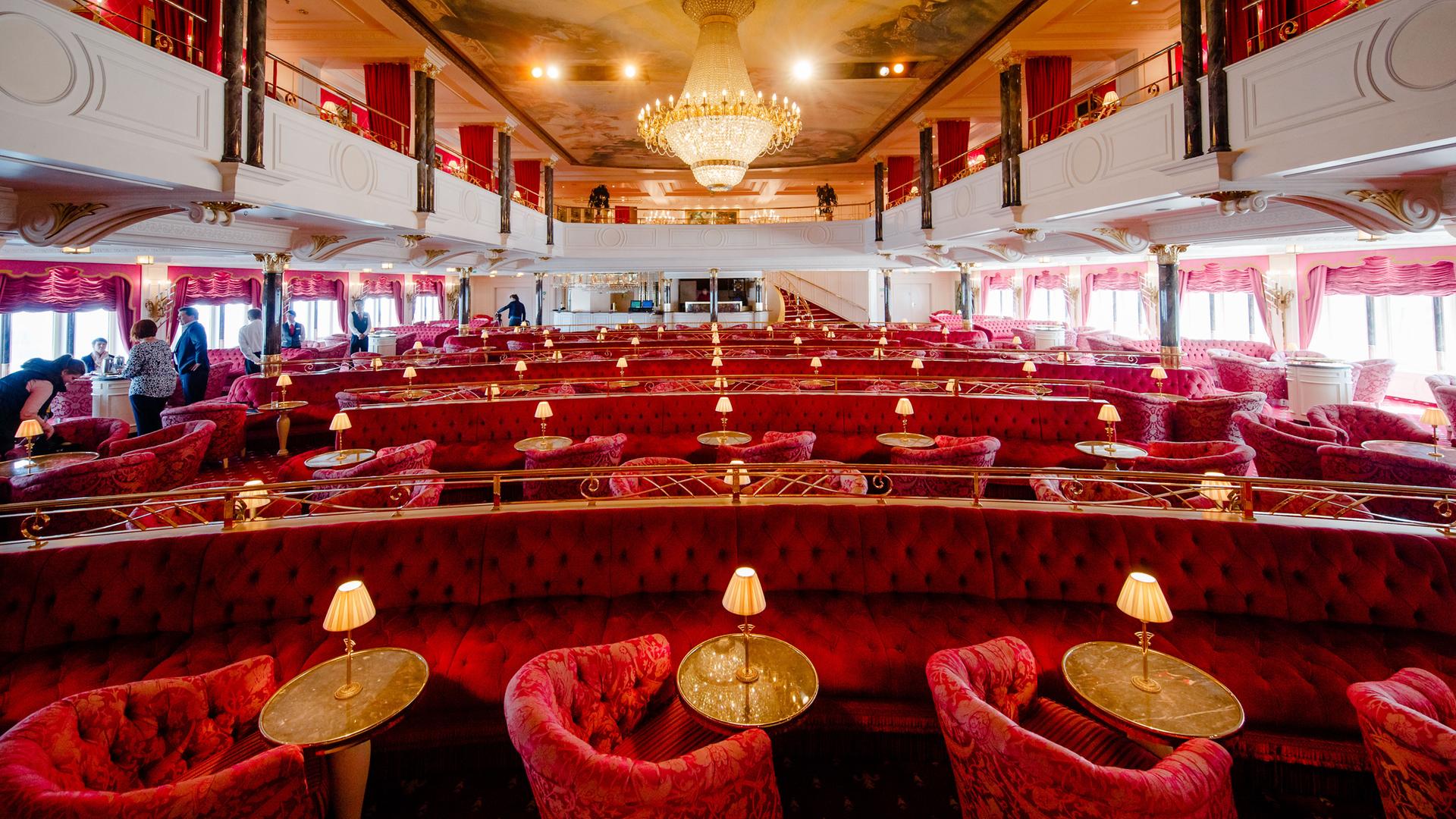 in Kaisersaal "MS Germany" Armchairs and benches covered in red velvet stand around the small tables.  A large chandelier hangs in the center of the hall.