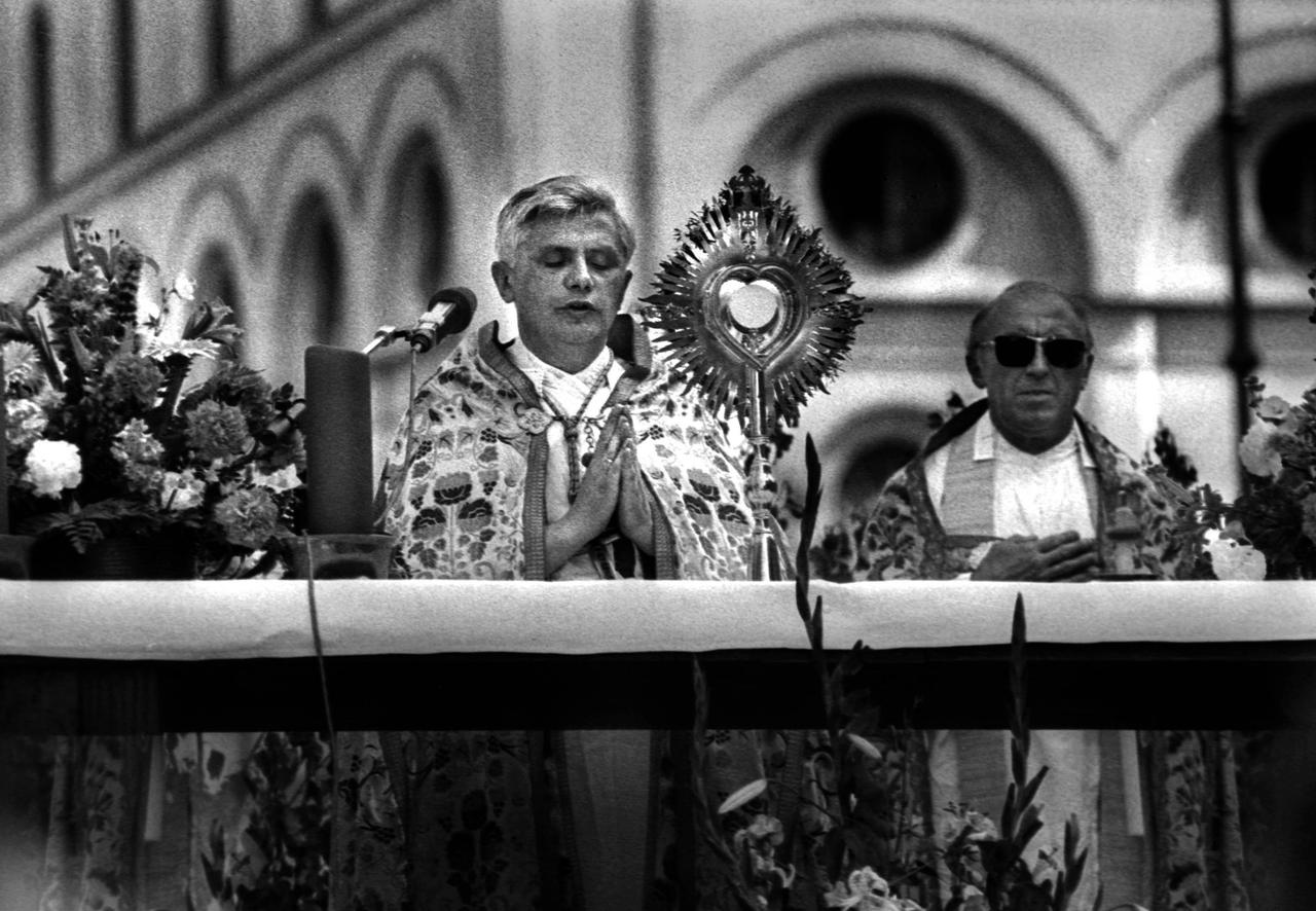 Schwarzweißfoto vom damaligen Erzbischof Joseph Ratzinger, der während einer Fronleichnamsprozession betend mit geschlossenen Augen in ein Mikrofon spricht.
