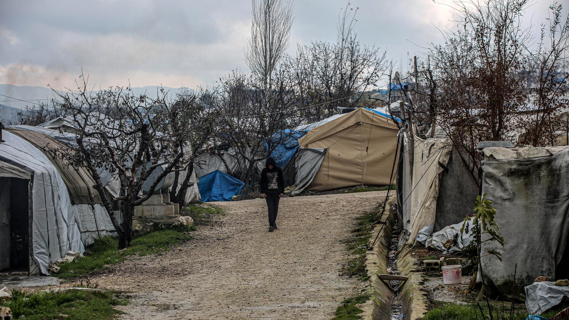 Eine Person mit hochgezogener Kapuze läuft eine unbefestigte Straße im Flüchtlingscamp entlang, die von Zelten gesäumt ist. Eine Person mit hochgezogener Kapuze läuft eine unbefestigte Straße im Flüchtlingscamp entlang, die von Zelten gesäumt ist.