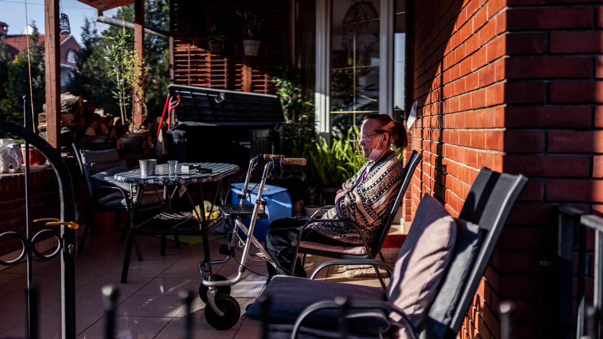 Eine ältere Frau sitzt auf der Terrasse eines Wohnhauses. Vor ihr steht ein Rollator.