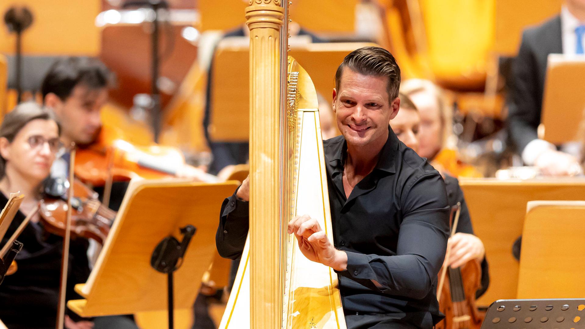 Ein Mann sitzt in schwarzer Konzertkleidung an einer goldenen Harfe. Die Hände an den Saiten. Er lächelt. Hinter ihm sind einzelne Orchestermusiker zu erkennen. Ein Mann sitzt in schwarzer Konzertkleidung an einer goldenen Harfe. Die Hände an den Saiten. Er lächelt. Hinter ihm sind einzelne Orchestermusiker zu erkennen.