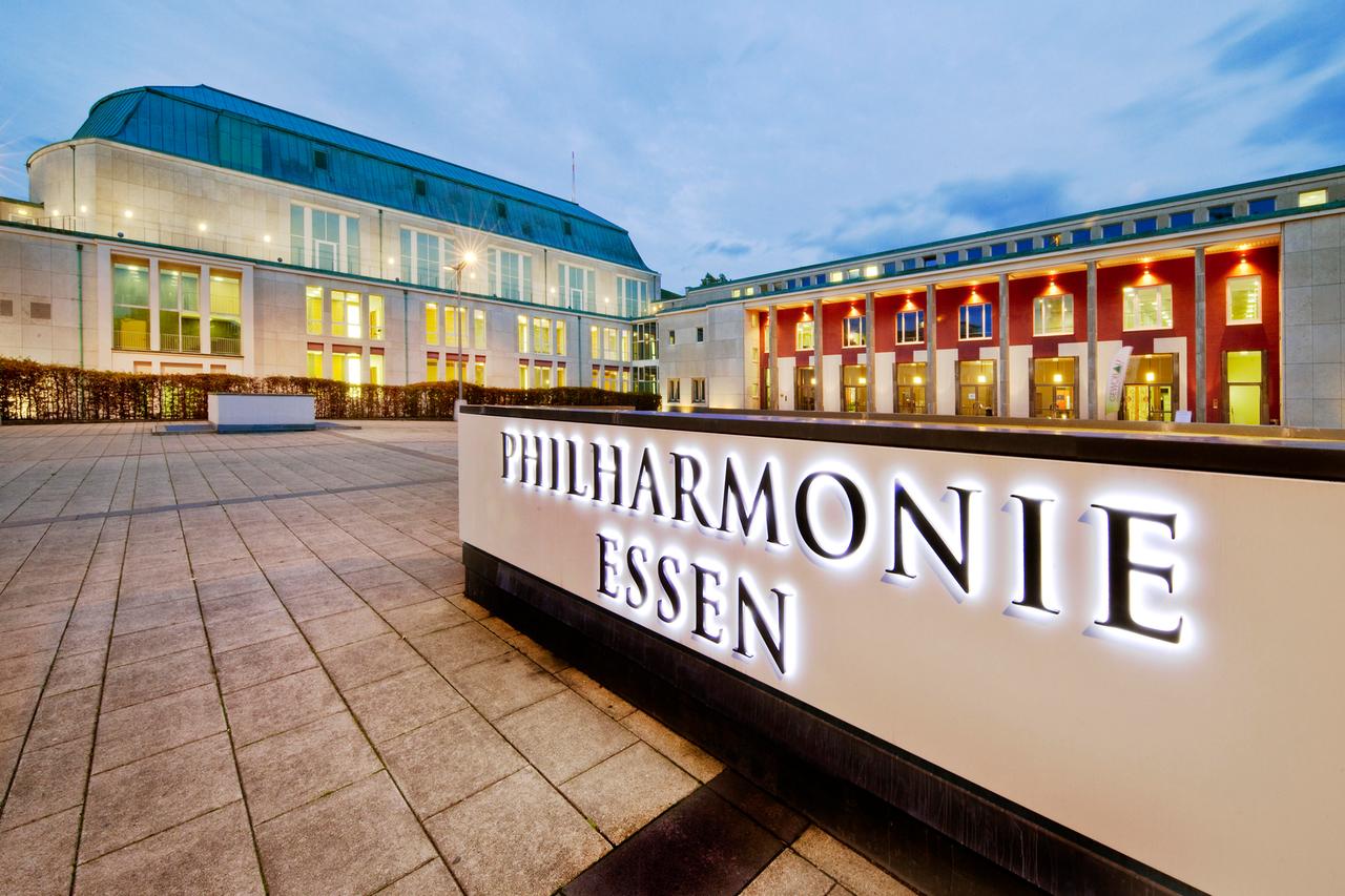 Im Vordergrund leuchtet der Schriftzug der Philharmonie Essen. Im Hintergrund steht das erleuchtete Konzerthaus. Im Vordergrund leuchtet der Schriftzug der Philharmonie Essen. Im Hintergrund steht das erleuchtete Konzerthaus.