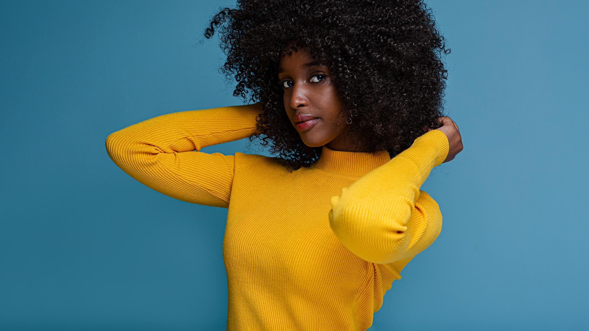 Eine weibliche Person of Color steht in gelbem Pullver vor einer blauen Wand.