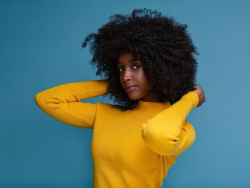 Eine weibliche Person of Color steht in gelbem Pullver vor einer blauen Wand.