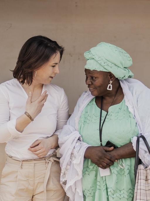 Die Bundesaussenministerin Annalena Bearbock steht mit einer Frau in traditioneller Tracht vor einer Hauswand, Mali 2022. 