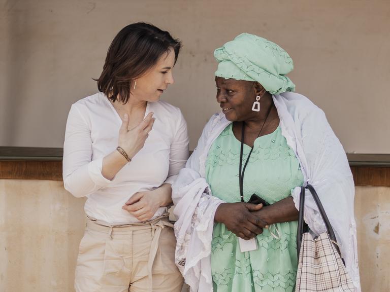 Die Bundesaussenministerin Annalena Bearbock steht mit einer Frau in traditioneller Tracht vor einer Hauswand, Mali 2022. 