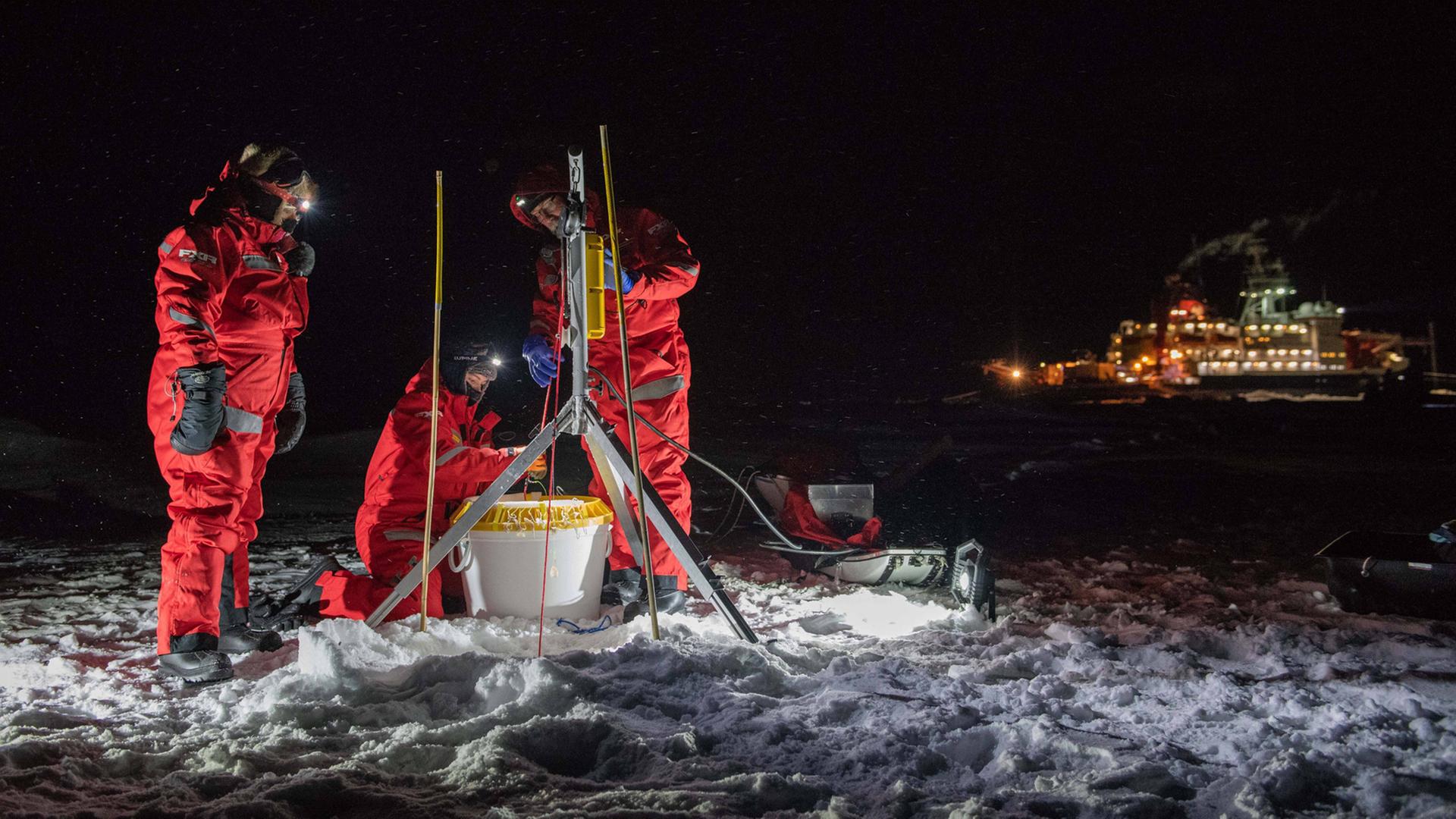 Drei Forschende entnehmen in der Polarnacht Proben an einem Loch im Eis, im Hintergrund die "Polarstern" Drei Forschende entnehmen in der Polarnacht Proben an einem Loch im Eis, im Hintergrund die "Polarstern"