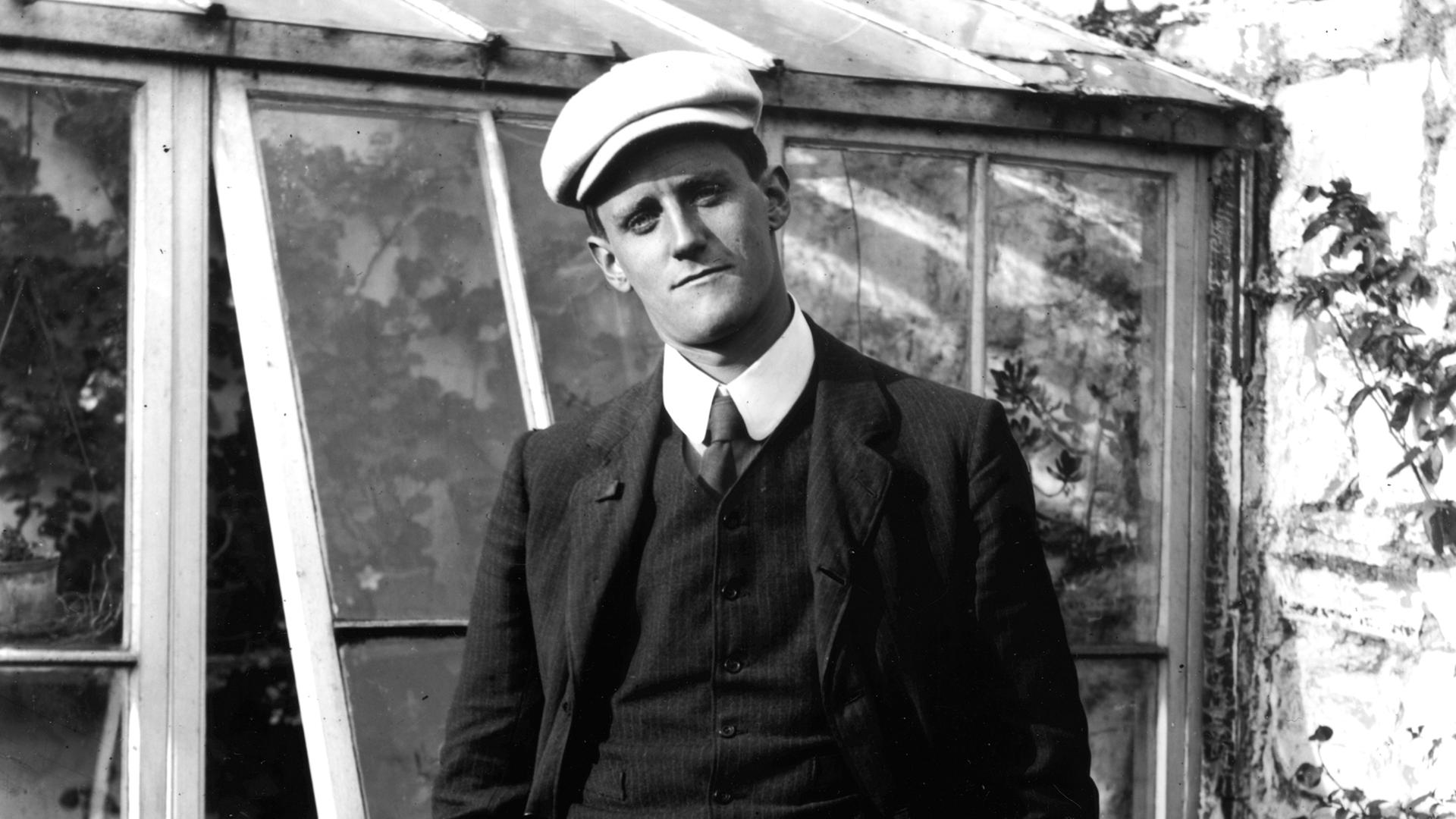 Der Schriftsteller James Joyce steht vor einem Gewächshaus, trägt Schiebermütze und schaut in die Kamera. Der Schriftsteller James Joyce steht vor einem Gewächshaus, trägt Schiebermütze und schaut in die Kamera.