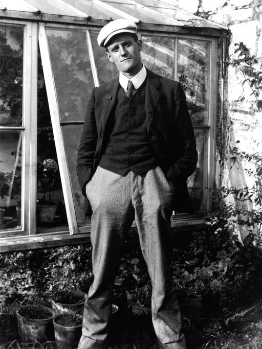 Der Schriftsteller James Joyce steht vor einem Gewächshaus, trägt Schiebermütze und schaut in die Kamera.
