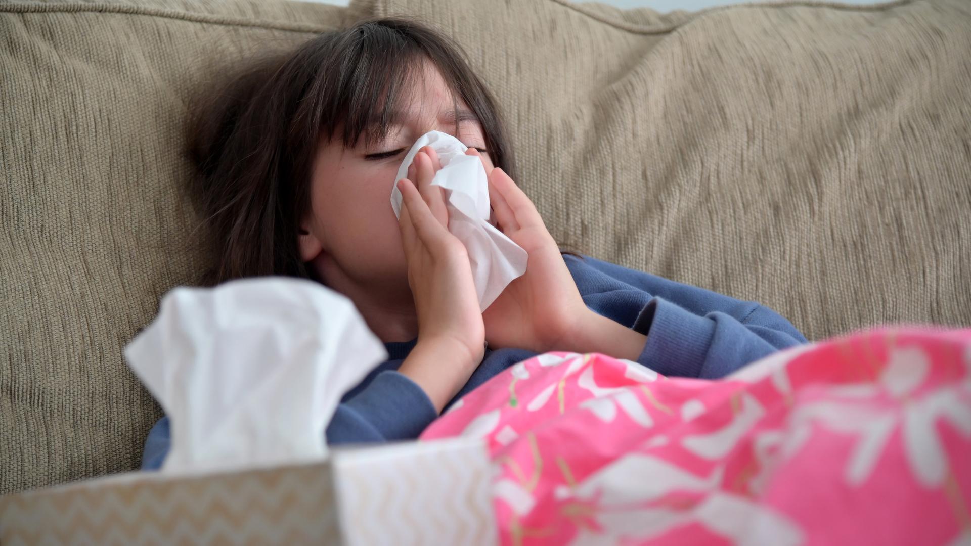 Ein Mädchen sitzt auf dem Sofa und putzt sich die Nase mit einem Taschentuch. 