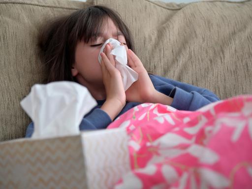 Ein Mädchen sitzt auf dem Sofa und schnäuzt sich die Nase. Wegen zunehmender Fälle von Infektionen mit dem Respiratorischen Synzytial-Virus (RSV) bei Kindern spitzt sich die Lage in vielen Kliniken zu