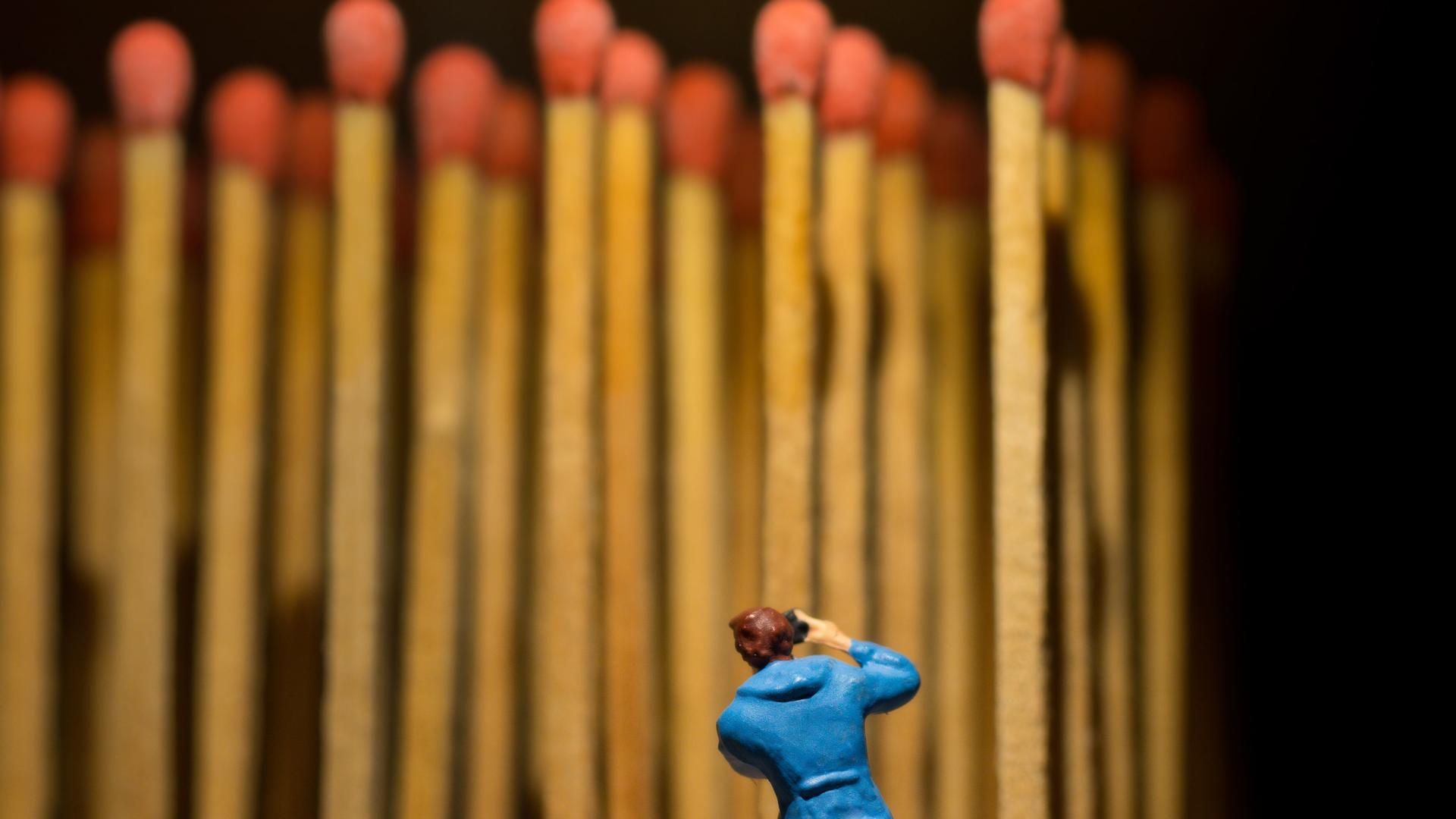 Eine Miniatur-Figur steht vor einer Reihe aufrecht stehender Streichhölzer mit roten Zündkopf. Eine Miniatur-Figur steht vor einer Reihe aufrecht stehender Streichhölzer mit roten Zündkopf.