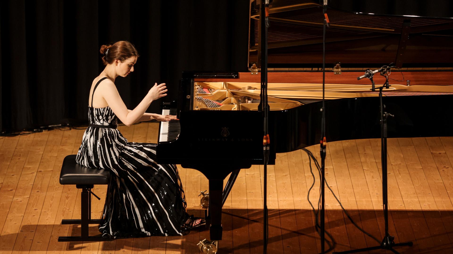 Eine junge Pianistin sitzt an einem schwarzen Konzertflügel. Eine Hand liegt auf den Tasten, die andere hebt sie gerade hoch. Sie trägt ein schwarz weiß gestreiftes Kleid und hat rotbraune Haare.