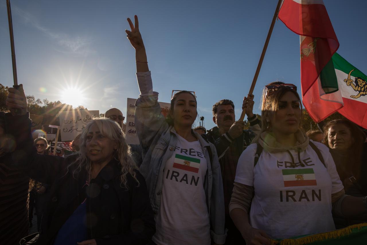 Demonstration aus Solidarität mit den Protestierenden im Iran. Im Vordergrund zwei Frauen mit T-Shirts auf denen steht: Free Iran. Dazwischen eine iranische Flagge. Ein Mann dahinter hält eine iranische Flagge hoch. Vor blauem Himmel im Hintergrund scheint die Sonne.