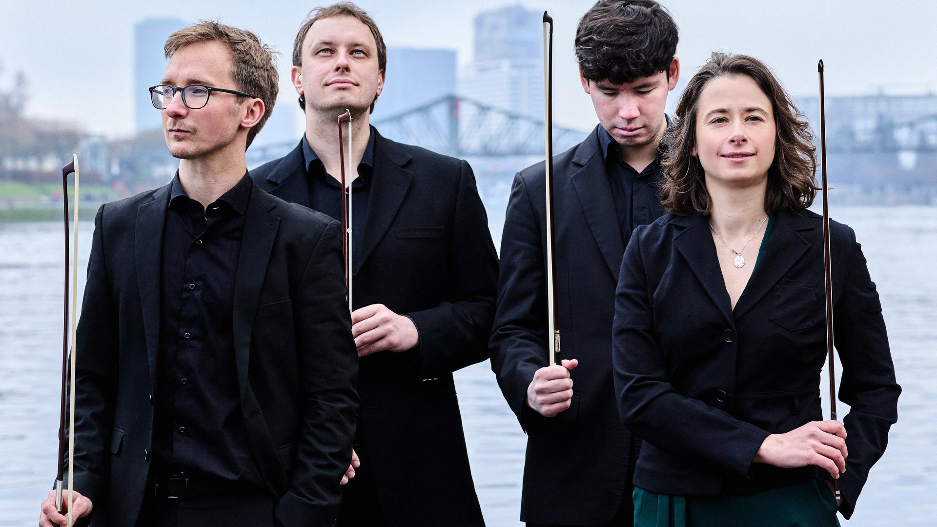 Vier junge Menschen stehen vor einem Stadtpanorama. Sie halten alle einen Bogen in der Hand, mit sie auf ihren Instrumenten spielen können. Die Instrumente: Geige, Bratsche, Cello sind aber nicht sichtbar. Zusammen bilden die vier das Eliot Quartett. Vier junge Menschen stehen vor einem Stadtpanorama. Sie halten alle einen Bogen in der Hand, mit sie auf ihren Instrumenten spielen können. Die Instrumente: Geige, Bratsche, Cello sind aber nicht sichtbar. Zusammen bilden die vier das Eliot Quartett.
