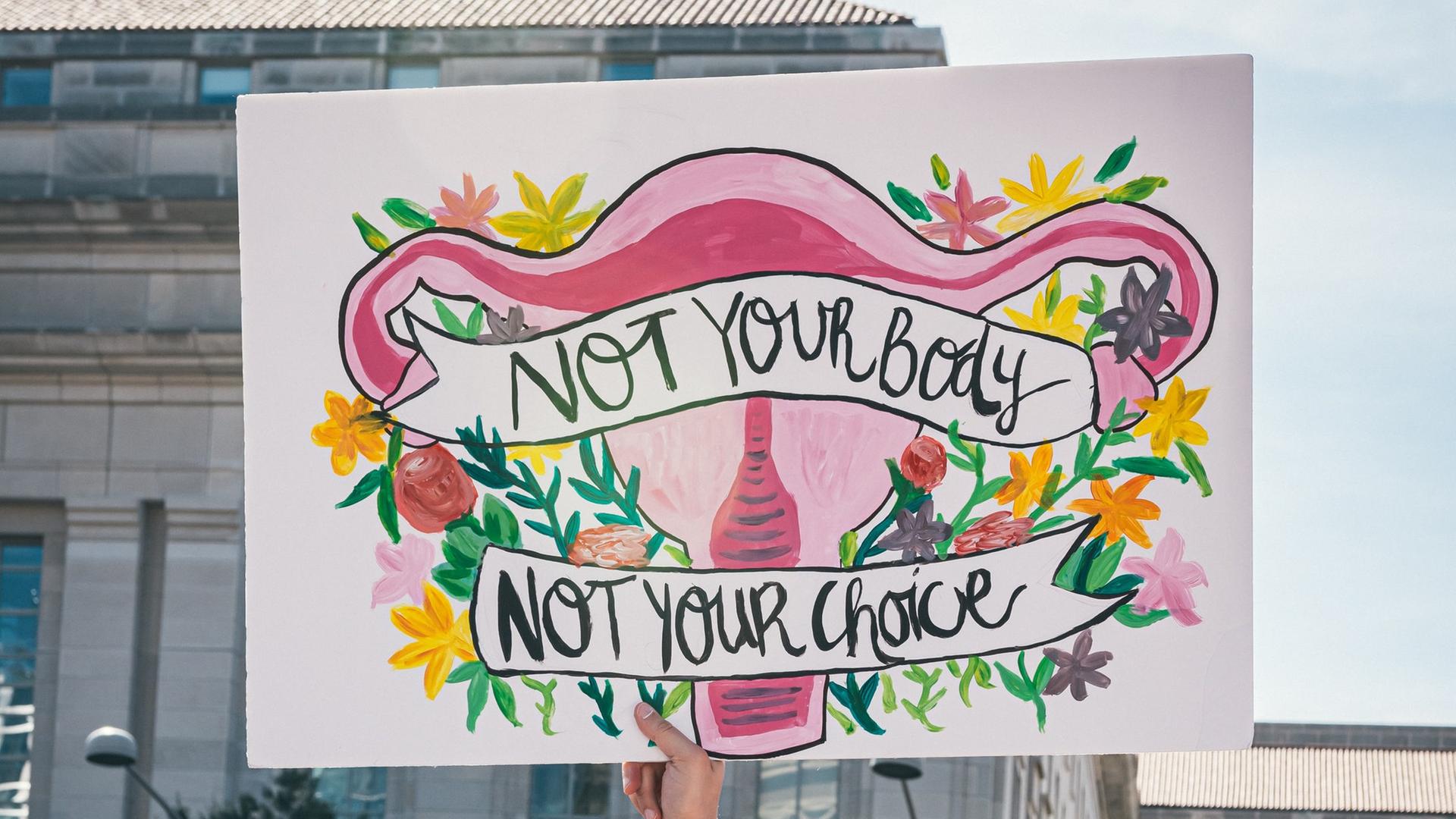 Auf einer Demonstration für Frauenrechte wird ein bunt bemaltes Plakat hochgehalten. Es zeigt die weiblichen Geschlechtsorgane umrahmt von Blumen mit der Aufschrift: "Not your body - not your choice".