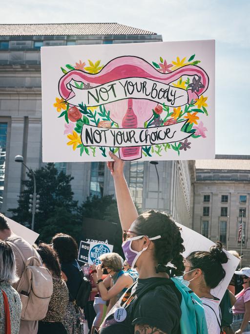 Auf einer Demonstration für Frauenrechte wird ein bunt bemaltes Plakat hochgehalten. Es zeigt die weiblichen Geschlechtsorgane umrahmt von Blumen mit der Aufschrift: "Not your body - not your choice".