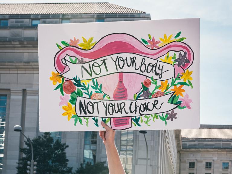 Auf einer Demonstration für Frauenrechte wird ein bunt bemaltes Plakat hochgehalten. Es zeigt die weiblichen Geschlechtsorgane umrahmt von Blumen mit der Aufschrift: "Not your body - not your choice".