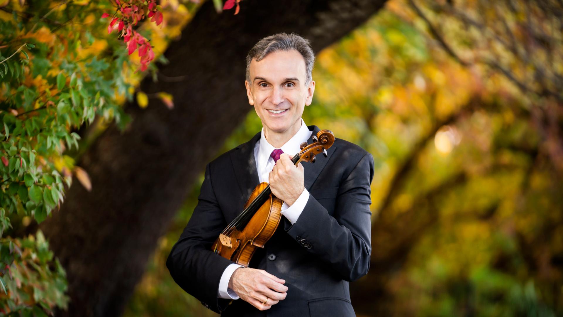 Der Geiger Gil Shaham steht zwischen üppigen Bäumen mit Anzug uns einer Geige.