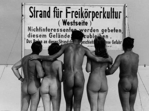 Eine Gruppe von Männern und Frauen steht  nackt mit dem Rücken zum Betrachter vor einem Schild "Strand für Freikörperkultur - Westseite".