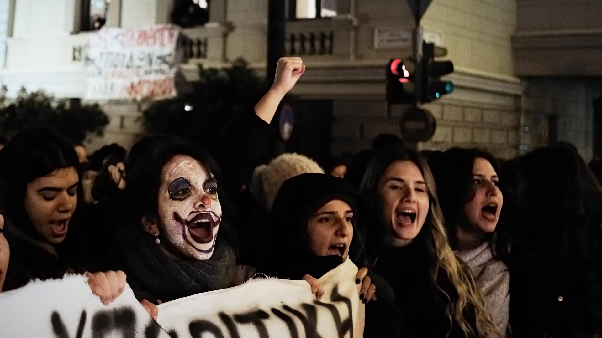Dunkel gekleidete junge Menschen protestieren in der Dunkelheit in Athen, in der Mitte eine junge Frau mit weiß geschminktem Clownsgesicht. Dunkel gekleidete junge Menschen protestieren in der Dunkelheit in Athen, in der Mitte eine junge Frau mit weiß geschminktem Clownsgesicht.
