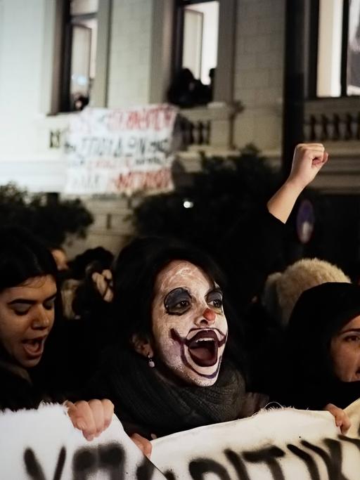 Dunkel gekleidete junge Menschen protestieren in der Dunkelheit in Athen, in der Mitte eine junge Frau mit weiß geschminktem Clownsgesicht.