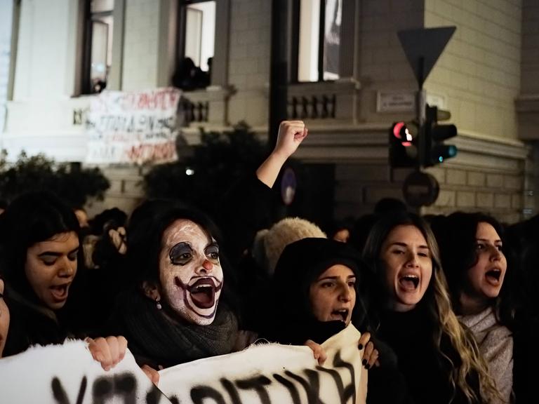Dunkel gekleidete junge Menschen protestieren in der Dunkelheit in Athen, in der Mitte eine junge Frau mit weiß geschminktem Clownsgesicht.