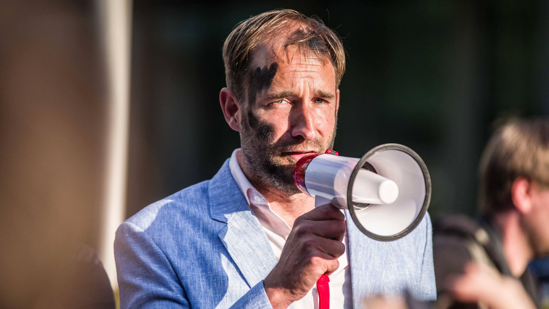 Der Aktionskünstler Philipp Ruch steht mit schwarz angemaltem Gesicht auf einer Straße und hält ein Megafon vor das Gesicht. Der Aktionskünstler Philipp Ruch steht mit schwarz angemaltem Gesicht auf einer Straße und hält ein Megafon vor das Gesicht.
