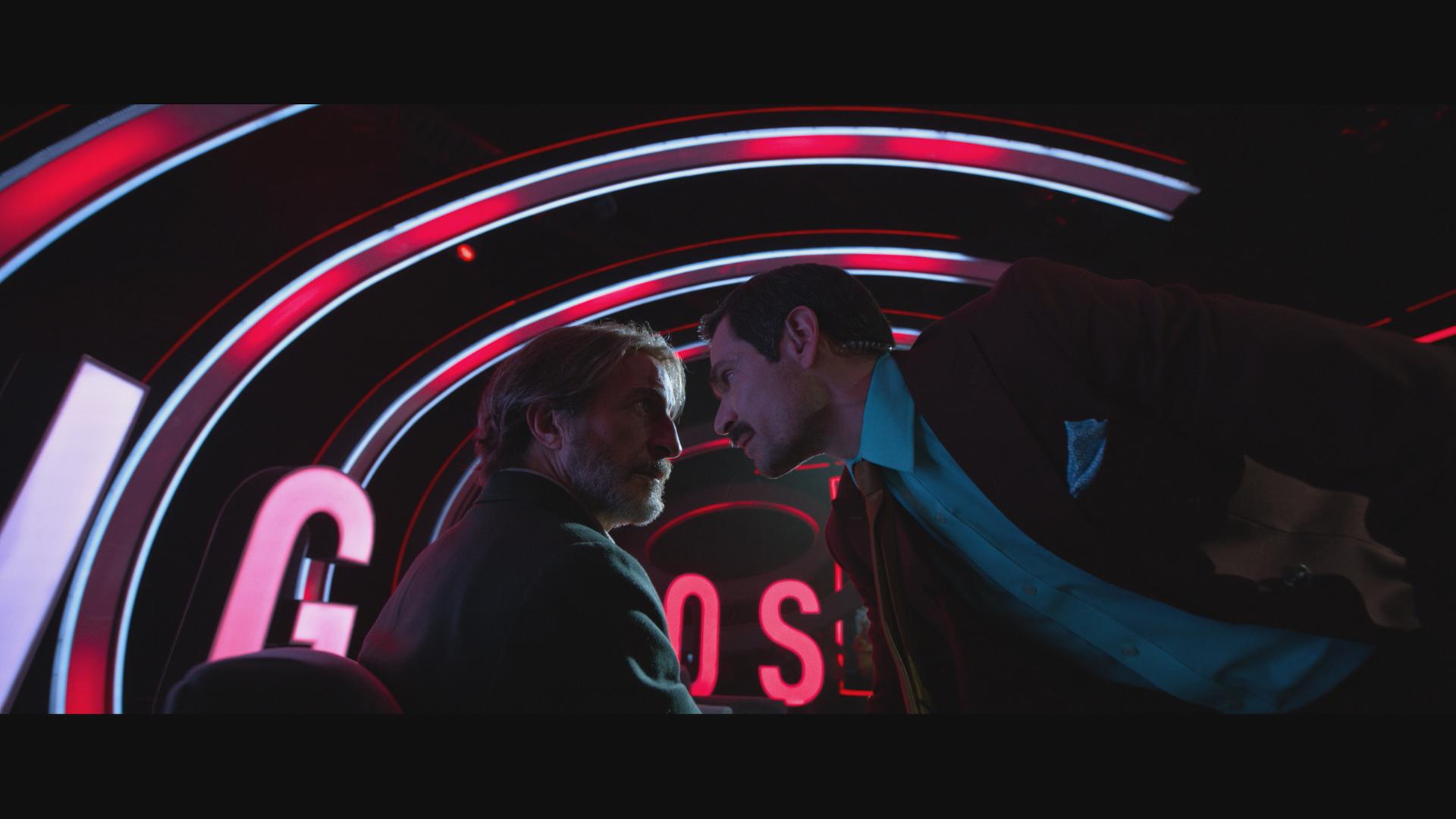 Ein Mann im Anzug schiebt sein Gesicht ganz nah an das Gesicht eines bärtigen Mannes. Im Hintegrund sind gebogene rote Leuchtstoffröhren zu sehen. 
