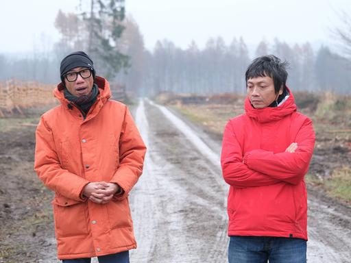 Iswanto Hartono und Reza Afisina von der Künstlergruppe Ruangrupa tragen orange Anoraks, hinter ihnen ist ein Forstweg zu sehen.