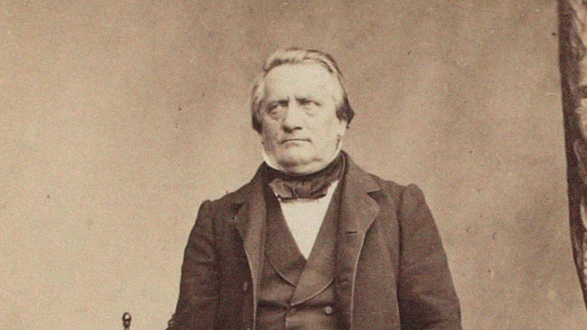 Ein schwarz weiß Foto des Komponisten Franz Lachner. Er steht vor einem Stuhl, lehnt mit der linken Hand auf einem Tisch. Er blickt etwas zur Seite.