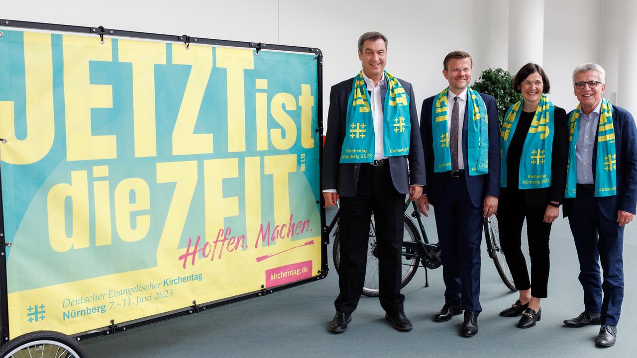 Neben einem Plakat mit dem Motto des Kirchentags ("Jetzt ist die Zeit") stehen Bayerns Ministerpräsident Markus Söder, Nürnbergs Oberbürgermeister Marcus König, die Generalsekretärin des Deutschen Evangelischen Kirchentags, Kristin Jahn, sowie der Präsident des Deutschen Evangelischen Kirchentags, Thomas de Maizière. Alle vier tragen grüne Schals, auf denen in gelber Schrift das Kirchentagsmotto eingestickt ist. 