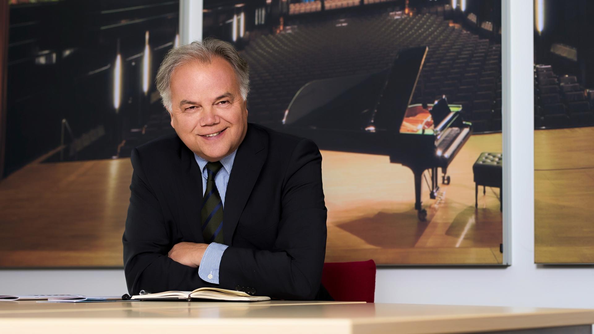 Ein Mann im Anzug sitzt mit verschränkten Armen an einem Schreibtisch vor einem großen Bild eines leeren Konzertsaales. Ein Mann im Anzug sitzt mit verschränkten Armen an einem Schreibtisch vor einem großen Bild eines leeren Konzertsaales.