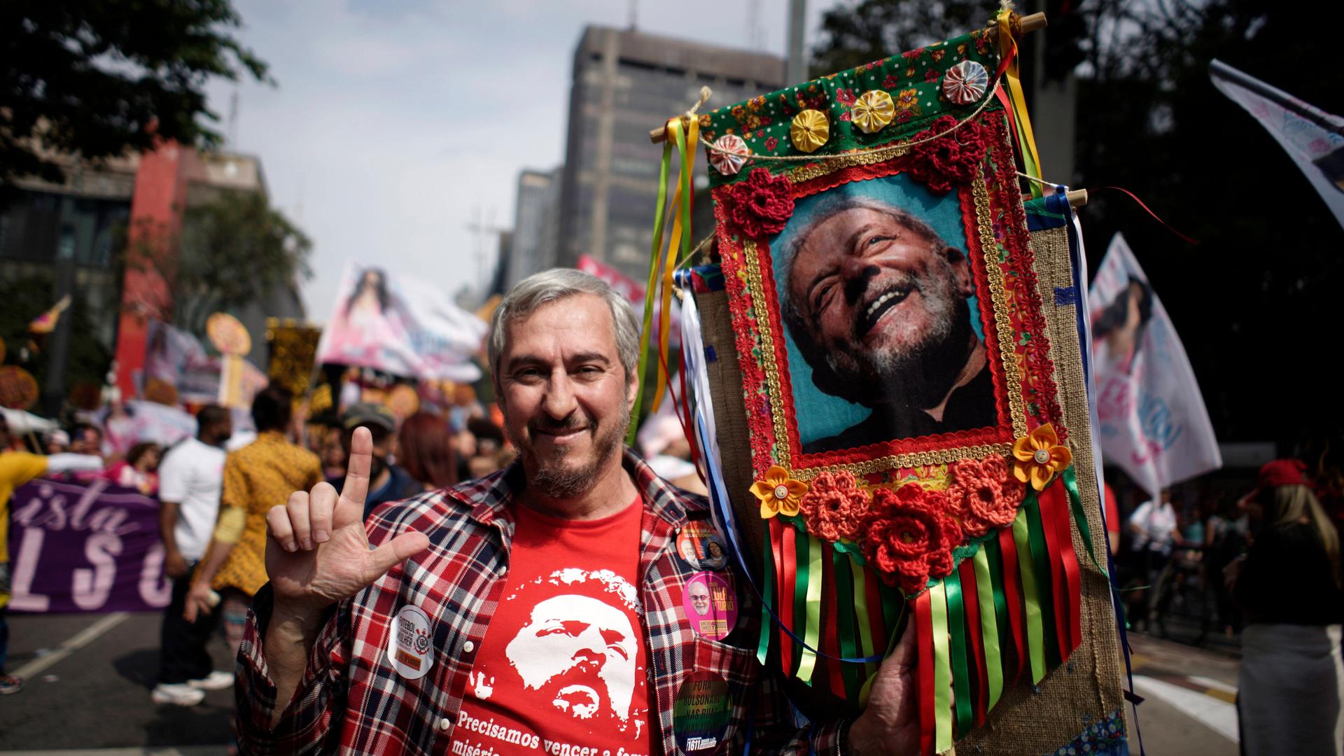 Ein Mann steht in einer Menge auf der Straße und hält ein großes buntumrandetes Foto von Lula da Silva an einem Stock in der linken Hand. Mit der rechten streckt er Zeigefinger und Daumen in die Höhe. Ein Mann steht in einer Menge auf der Straße und hält ein großes buntumrandetes Foto von Lula da Silva an einem Stock in der linken Hand. Mit der rechten streckt er Zeigefinger und Daumen in die Höhe.