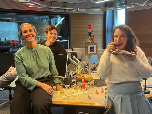 Sonja Koppitz, Utz Dräger und Caro Korneli sitzen auf dem Studiotisch. In der Mitte stehen Bildschirme und Mikrofone. Caro Korneli beißt in ein Stück Kuchen.