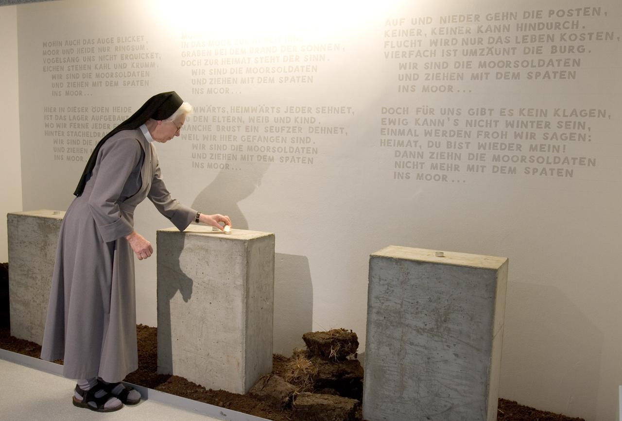 Eine Ordensschwester zündet in einem Gedenkraum eine Kerze an. Der schlichte Raum ist mit Beton-Stelen und Torf-Ballen eingerichtet, die Wand dahinter trägt eine Inschrift.