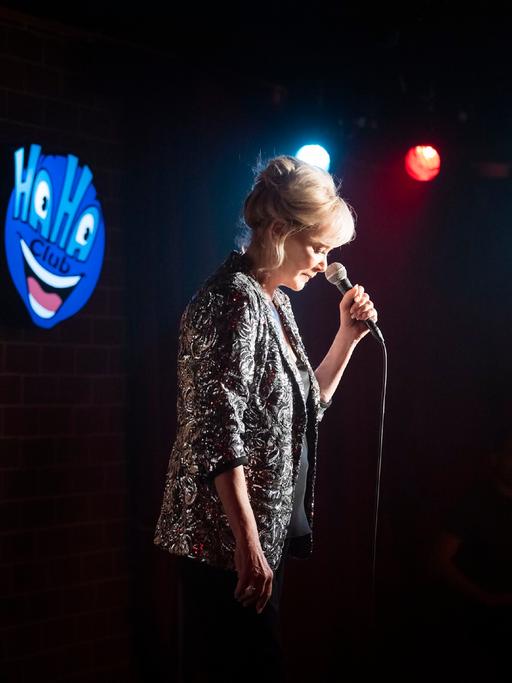 Filmszene aus der Serie "Hacks" (RTL +): Eine Frau im Spotlight gespielt von Jean Smart steht vor einem Mikrofon auf einer dunklen Bühne. Auf einem Schild steht Haha Club.