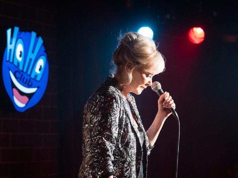 Filmszene aus der Serie "Hacks" (RTL +): Eine Frau im Spotlight gespielt von Jean Smart steht vor einem Mikrofon auf einer dunklen Bühne. Auf einem Schild steht Haha Club.