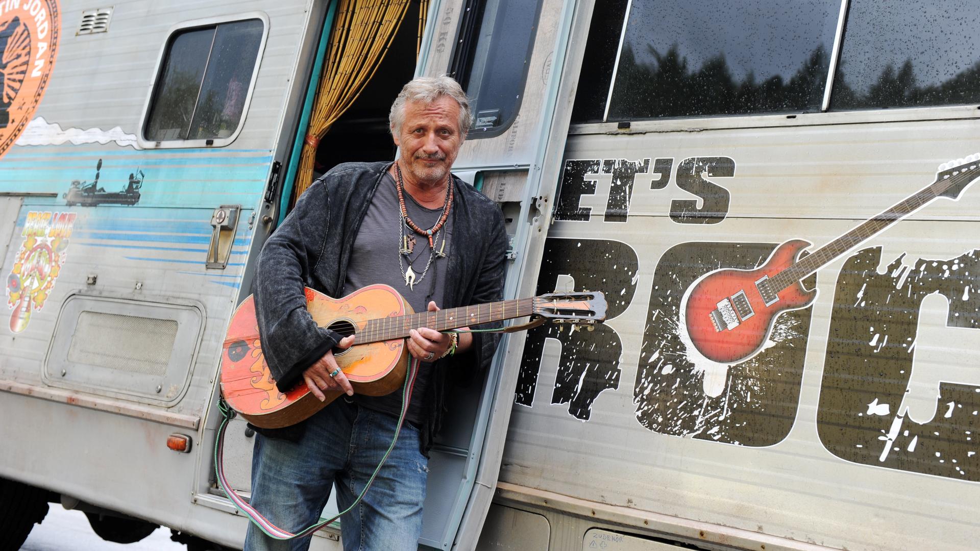 Ein Mann lehnt mit seiner Gitarre im Arm an einem Wohnwagen und lächelt in die Kamera. Es ist der Musiker Konstantin Wecker. 