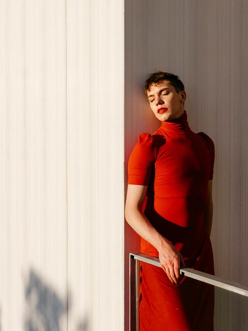 Ein Transgender Fluid Mann im roten Kleid und rotem Lippenstift steht im Sonnenlicht an eine Wand gelehnt.