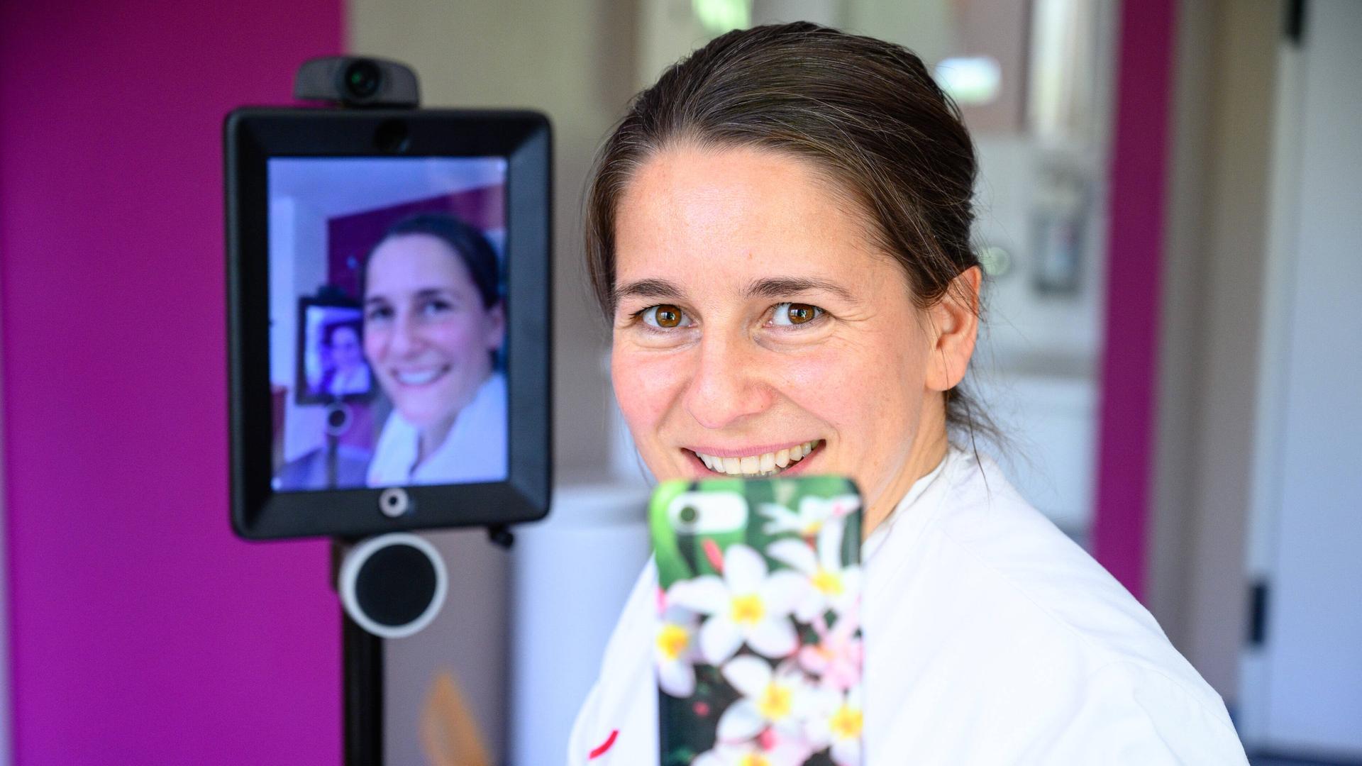 Die Gynäkologin Mandy Mangler hält ein Smartphone in der Hand. Hinter ihr sieht man ihr Gesicht auf einem Monitor. Die Gynäkologin Mandy Mangler hält ein Smartphone in der Hand. Hinter ihr sieht man ihr Gesicht auf einem Monitor.