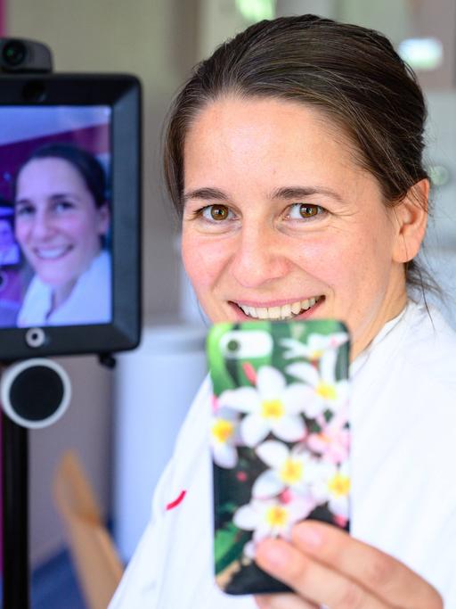 Die Gynäkologin Mandy Mangler hält ein Smartphone in der Hand. Hinter ihr sieht man ihr Gesicht auf einem Monitor.