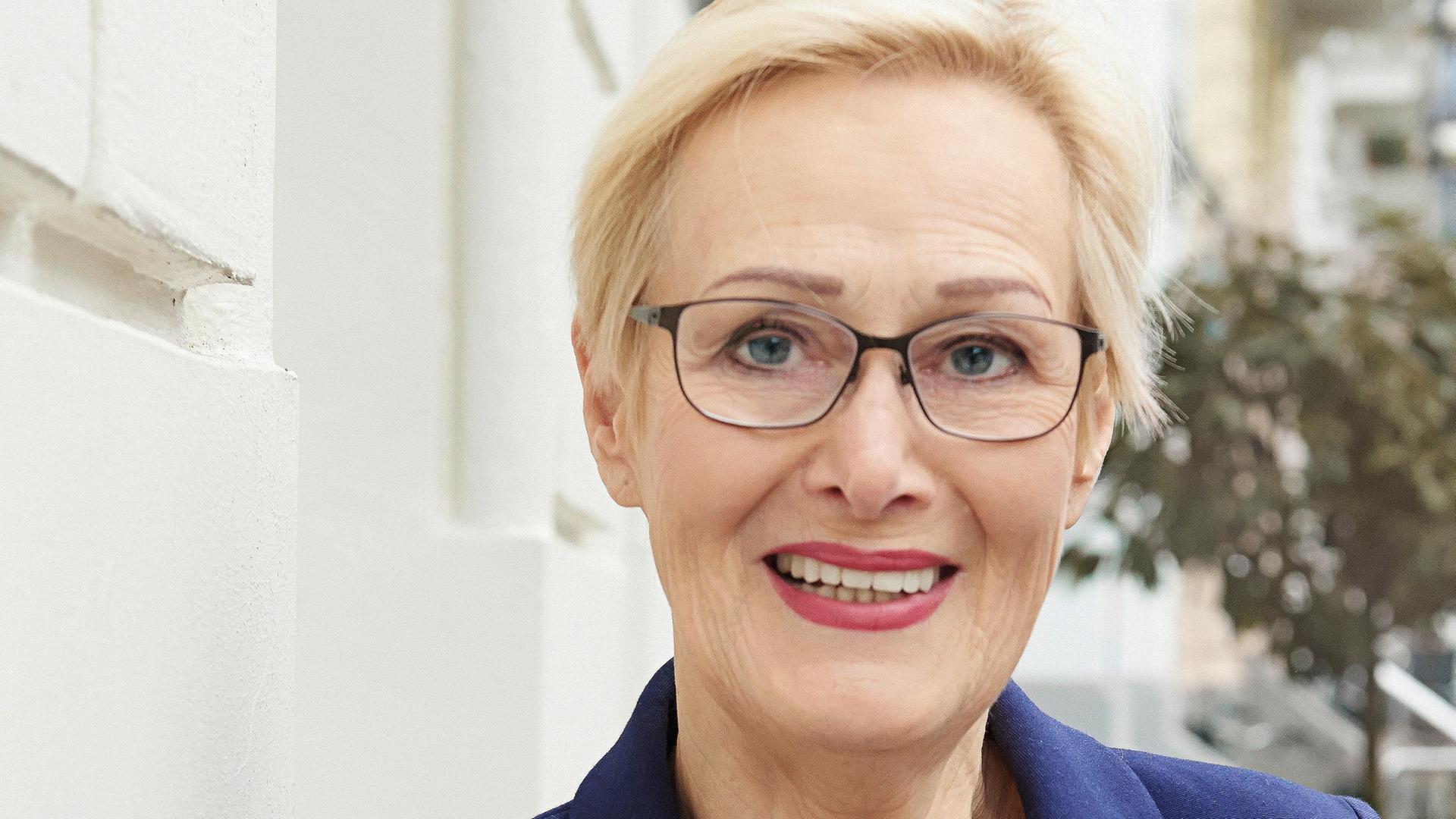 Porträt von Eva Wlodarek. Sie lehnt an einer hellen Hauswand und trägt einen blauen Blazer und einen gestreiften Pullover. Porträt von Eva Wlodarek. Sie lehnt an einer hellen Hauswand und trägt einen blauen Blazer und einen gestreiften Pullover.