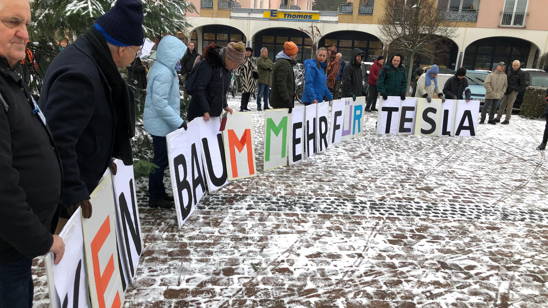 Menschen stehen im Halbkreis auf einem gepflasterten Platz. Sie halten Transparente mit einzelnen Buchstaben vor sich, von links nach rechts kann man lesen: "Kein Baum mehr für Tesla" Menschen stehen im Halbkreis auf einem gepflasterten Platz. Sie halten Transparente mit einzelnen Buchstaben vor sich, von links nach rechts kann man lesen: "Kein Baum mehr für Tesla"