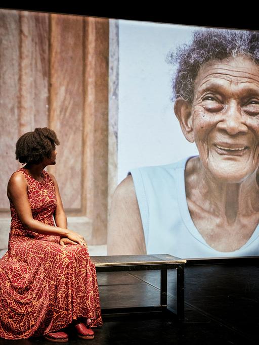 Eine junge schwarze Frau in einem roten Kleid sitzt auf einer Bank auf einer Theaterbühne und schaut auf eine Leinwand, auf der eine alte schwarze Frau abgebildet ist.